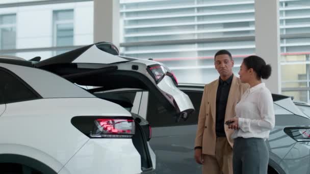 アフリカ系アメリカ人自動車ディーラーの販売店の女性はショールームのクライアントビジネスマンバイヤーが電気自動車のクロスオーバー現代技術の自動車の特徴の新しい機能を開く自動トランクを示すのを助けます — ストック動画