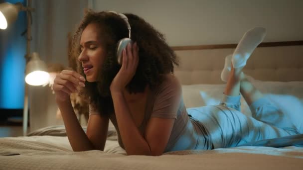 リラックス幸せなアフリカ系アメリカ人のエスニックガールは ワイヤレスイヤホン若い女性の居心地の良いベッドの上に曲を歌います ジーンエイジャー リラックスした夜の家で音楽を聞く ヘッドフォン携帯電話の休日を楽しむ — ストック動画