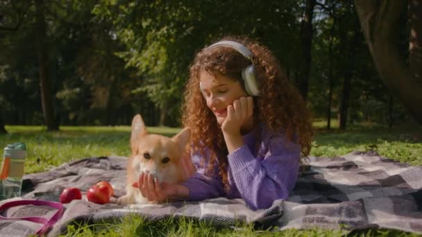 ヘッドフォンガールの白人若い女性幸せな女性ペットの飼い主は草の自然にかわいい小さなウェールズのコルギの子犬を飼っています 笑顔の女性ハンドラーは公園で家畜の犬のトリートを飼います — ストック動画