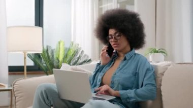 Afro-Amerikalı kadın etnik bayan kanepe cevap veriyor. Kanepe başında bilgisayarla konuşan dizüstü bilgisayarını arıyor. İnternetten cep telefonu görüşmesi sipariş ediyor.