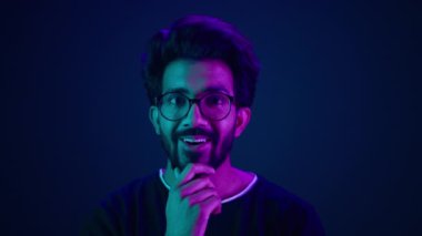 Şaşırmış, mutlu, şaşkın, şoke olmuş Arap, Hintli, erkek, programcı, internet geliştiricisi, iş adamı, yüzü morumsu, neon arka planda gülümseyen çeneye bakıyor.