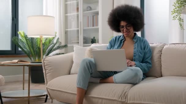 笑顔のアフリカ系アメリカ人女性 家庭でソファーで笑顔 ラップトップ笑顔のオンライン仕事 タイピング メッセージ 幸せな女性 コンピュータでチャット インターネットソーシャルメディア 面白いビデオを見る — ストック動画