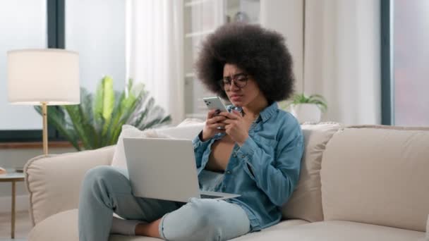 携帯電話のソファーにアフリカ系アメリカ人女性 女性学生フリーランサー ユーザーガジェット中毒インターネットソーシャルメディア中毒女性 スマートフォンを見ているラップトップを保持 — ストック動画