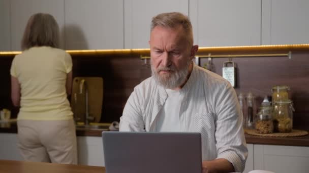 中年退職 50代男性夫灰色髪のビジネスマン コンピュータのノートブックと遠くで働くことを考える思慮深い問題解決のラップトップは家庭で働きますが 女性の妻は台所で料理します — ストック動画