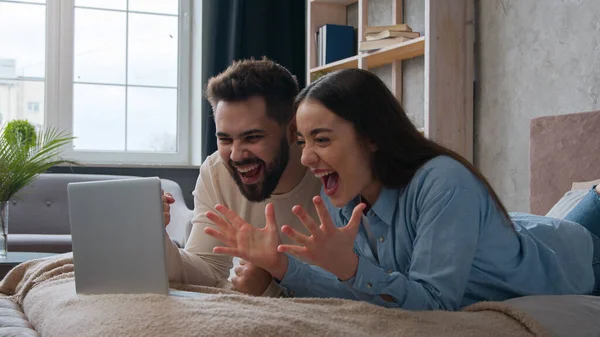 Excited Caucasian married couple lying on bed at home win laptop achievement read amazing news online bet victory celebrate success happy woman wife and man husband scream shout good result winning