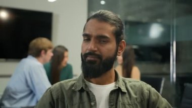 Arap kökenli Hintli etnik iş adamı müslüman erkek ofis yöneticisi profesyonel işadamı CEO 'su kameraya bakıyor arka plandaki bulanık takım arkadaşlarının masasında oturuyor