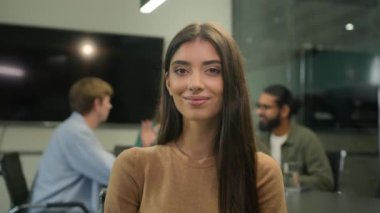 Beyaz kadın portresi, beyaz iş kadını, İspanyol kadın iş kadını, bayan yönetici, mutlu bir şekilde gülümseyen, kameraya bakan, ofisin arka planında bulanık iş arkadaşları, beşlik çakıyor.