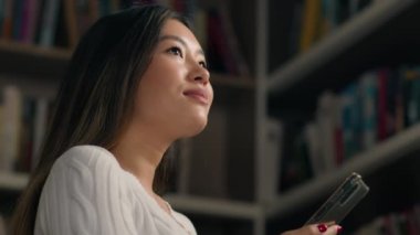 Üniversite kütüphanesinde kitap arayan Asyalı kadın etnik kız öğrenci telefon görev listesini kullanarak akıllı telefon modern teknoloji eğitiminde cep telefonu kullanımını kontrol ediyor.