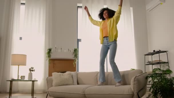 活跃有趣的疯狂的非洲裔美国女孩快乐的女人在客厅的沙发上跳起舞兴奋的女人跳起舞大声的音乐回家派对快乐的无忧无虑的快乐的女人周末度假自由 — 图库视频影像