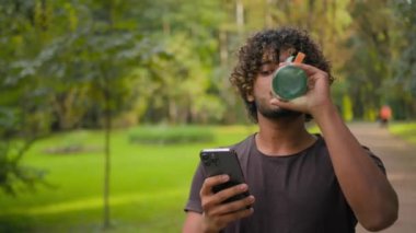 Arap Müslüman, Hintli, parkta akıllı telefonuyla yürüyen, spor şişesinden su içen, açık havada koşan, mutlu, gülümseyen erkek, cep telefonu uygulamasındaki antreman hedeflerini kontrol eden, etnik susamış adam.