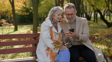 Kafkasyalı emekli çift aşık olgun kadın erkek banka oturup cep telefonu aygıtlarıyla gülümseyen eşler eşleri ile birlikte şehirde yaşlılık haberleri ve akıllı telefon okuyorlar.