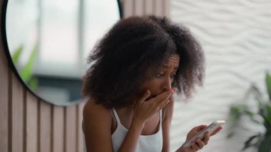 Afro-Amerikalı, banyo banyosundaki hoşnutsuz genç kadın, akıllı telefon ekranına bakıyor. Kötü haber spam dolandırıcılığı, etnik melez kız, mutsuz, cep telefonu hatasını vurguladı.