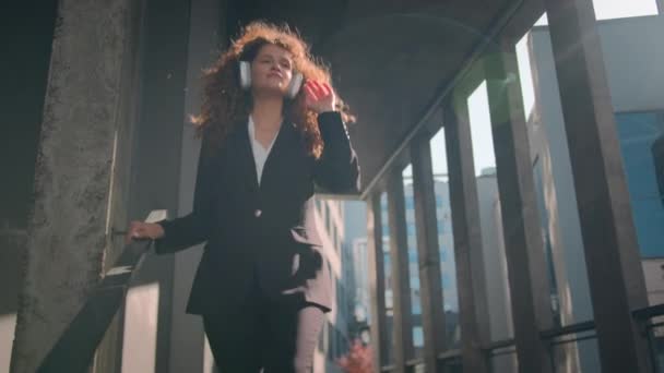 ハッピーガール コーカサス ビジネス 面白い ケアフリーダンス アウトドア ビジネスウーマン ダウン オフィスビルの階段を歩く ヘッドフォンの自由の音楽を聴く — ストック動画