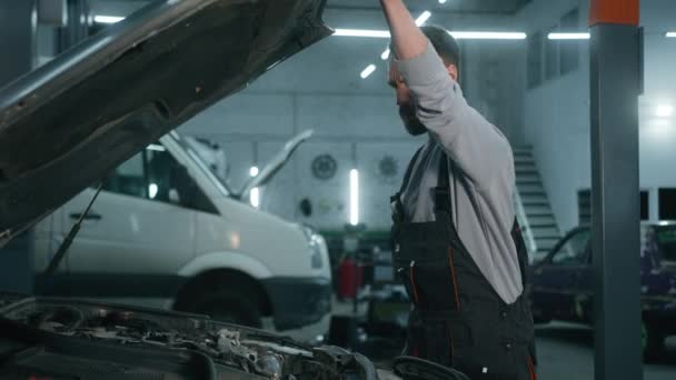 混淆不清的白人男子汽车修理工敞篷帽修理坏了的汽车在加油站擦着头汽车撞毁了汽车修理厂修理车辆 — 图库视频影像