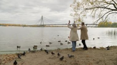 Kafkasyalı orta yaşlı çift mutlu kadın ve erkek sonbahar parkında gölde ördek besliyor iki emekli komik yaşlı insan kuşlarla el sallıyor ve tatillerde eğleniyorlar.