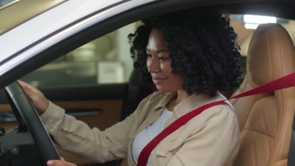 幸せなアフリカ系アメリカ人女性は車の笑顔の女の子バイヤーの車の内側の運転席に座ってステアリングホイールを回して屋内でディーラーで自動車のテストドライブを選択する自動車ショールームで新しい輸送を購入することを夢見ています — ストック動画