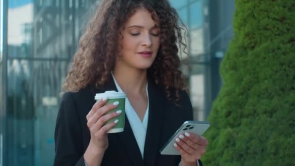 自信的白种人女人在纸杯里喝咖啡欧洲20多岁的女孩走在城市外面微笑的商人女人聊天发短信上网浏览智能手机小工具在城市里的商业网络 — 图库视频影像