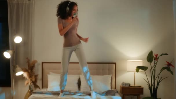 非洲裔美国女人积极快乐有趣20多岁的年轻女性晚上在家里的床上跳起舞 卧室里的傻女孩戴着耳机听音乐 用手机跳舞 心情很开心 — 图库视频影像