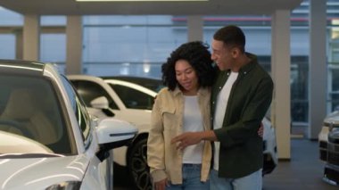 Afro-Amerikalı çift, galeri otomobil galerisinde modern çapraz araba arıyor. Erkek müşteriler, araç kuaföründe yürüyen müşteriler, birlikte yeni ve pahalı otomobil alımları seçmeyi tartışıyorlar.
