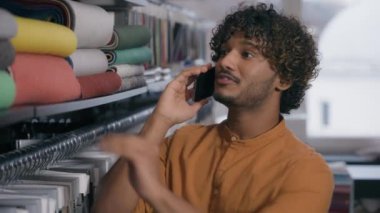 Kumaş koleksiyonu olan mutlu Hintli adam müşterisi kumaş ipeği kumaş ipeği kumaş kumaş örnekleri seçiyor gülümseyen adam kumaş dükkanında cep telefonu rengini tartışıyor.