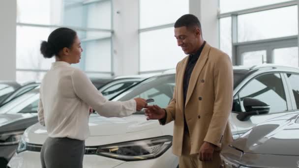 自動車ディーラーの販売代理店のマネージャーの女性はディーラーの中心の幸せな所有者アフリカのアメリカ人の男に車のキーを与えます 販売の顧客の賃料は新しい高級自動車の点検現代電気自動車警報システムを買います — ストック動画