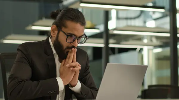 Pensive thinking business man Indian gut Arabian businessman boss leader employer male Muslim entrepreneur working with laptop think thoughtful rubbing hands ponder idea computer task wish in office