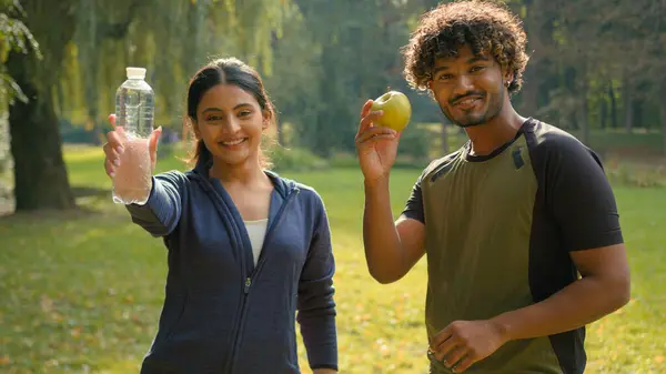 Indian man and woman relaxing after sports together in park outdoors Arabian fitness couple friends in city sport happy people showing holding apple fresh healthy fruit and bottle of water lifestyle