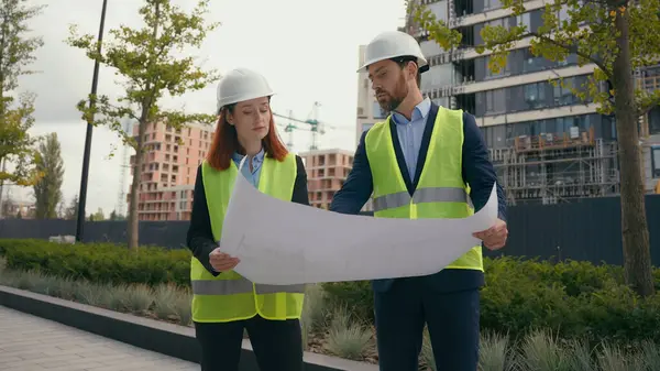 Two people workers builders in safety vests uniform hard hats discuss blueprint document paper plan of unfinished construction site discussion architects contractors man woman talk in city civil build