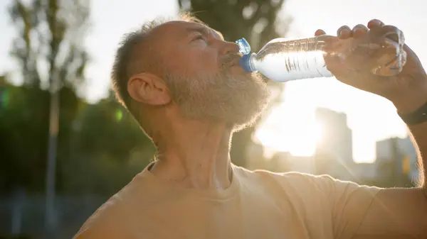 Close up older man drinking clear mineral water morning sun rays thirsty male drink pure aqua bottle city sunlight resting outside in park sport active sportsman healthy refreshing wellbeing energy