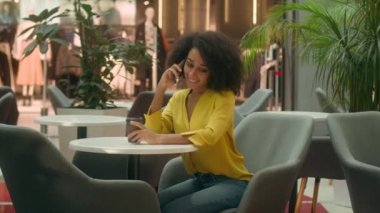 Genç Afrikalı Amerikalı kadın kafede oturup kahve içiyor cep telefonuyla konuşuyor cep telefonlarıyla sohbet ediyor alışveriş merkezini arıyor hafta sonu dinleniyor gülümseyen kadın iletişimi kuruyor zarifçe konuşuyor.