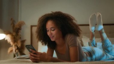 Afro-Amerikan etnik kadın 20 'li yaşlarda kıvırcık saçlı, cep telefonu kıvırcık, uykusuzluk çekiyor, gülümsüyor, akıllı telefon cihazıyla yatakta yatan bir kadını hayal ediyor ve internetten flört uygulaması ile sohbet ediyor.