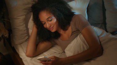 Rahat kız Afrikalı Amerikalı kadın, yatak odasında uyuyan bağımlı kadın gen z kaydırmalı internet uygulamaları kullanmadan önce uykusuzluk çekerek akıllı telefon sosyal medyasını tarıyor.