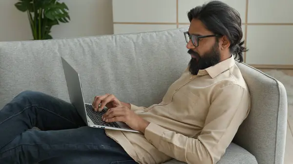 在家里工作的阿拉伯穆斯林自由职业者以千年为重点的印度商人在电脑上打字上网看屏幕躺在沙发上远程工作被隔离 — 图库照片