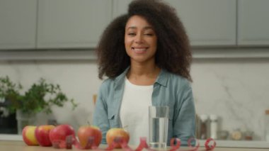 Gülümseyen Afro-Amerikan mutlu kadın etnik kız melez kadın ev mutfağında meyve elması bardak su ile gülümsüyor masa ağırlığı kontrol diyetinde bant sağlıklı beslenme