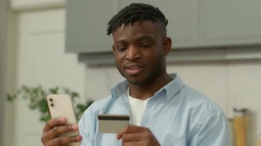 İnternet üzerinden telefon ve kredi kartı alışverişi yapan mutlu Afrikalı Amerikalı adam uzak mesafeden akıllı telefon alan, etnik erkek müşteri bankacılığı yapan, mutfaktan yemek siparişi veriyor.