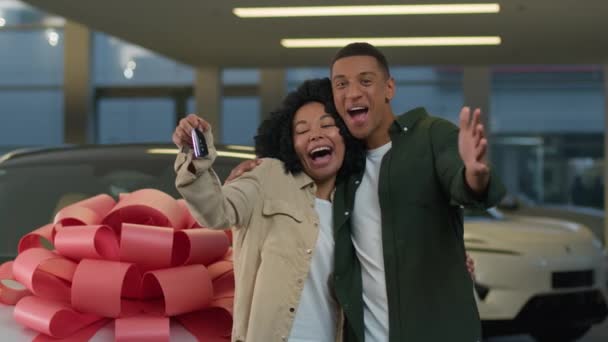 在现代经销商的陈列室里 兴奋的年轻夫妇欢欢喜喜地买新车 奢华的礼物 头戴大蝴蝶结的非洲裔美国女人男人展示着买汽车的钥匙 — 图库视频影像