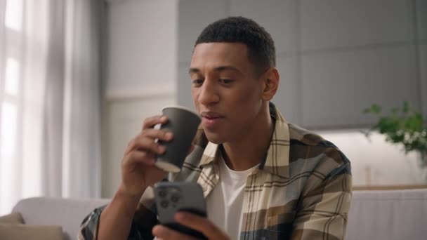 ハッピー笑顔のアフリカ系アメリカ人男性ユーザー ソファーの家でリラックスするソファーでエスニック男性 携帯電話を使用して喫茶コーヒー飲料スマイルチャットスマートフォンデバイス オンラインショッピングソーシャルメディア — ストック動画