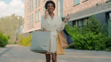 Mutlu gülümseme memnun gülümseme Afro-Amerikan kadın çift ırklı stil moda müşteri alıcısı alışveriş torbalarıyla şehirde yürüyor pozitif konuşan cep telefonu araması Kara Cuma