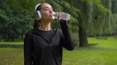 Güzel beyaz kadın fitness kızı, şehir parkındaki açık hava sporlarında gülümsüyor müzik kulaklıkları dinliyor, su içiyor, sağlıklı yaşam tarzı, ferahlatıcı, susuzluk giderici su içiyor.