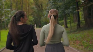 Arka planda iki beyaz kız aktif sporcu kadın mutlu bayan arkadaşlar parkta birlikte yürüyorlar spor antrenmanından sonra gülen kadınlar beşlik bir dostluk sarılması yapıyorlar.