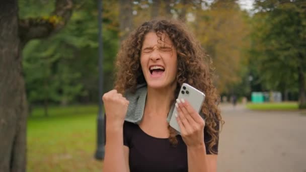 ハッピー驚いた驚いた驚きの女性コーカサスガールは携帯電話の読書ニュースの勝利を保持します スマートフォンショックで勝利を祝います 成功は都市公園のスポーツアウトドアの後に祝います — ストック動画