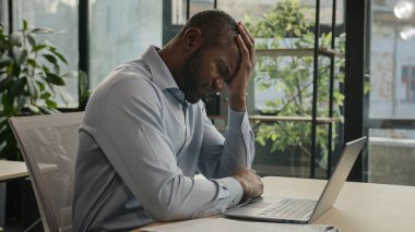Depresyondaki Afrikalı Amerikalı yetişkin iş adamı. Stresli hasta adam ofiste dizüstü bilgisayarla oturuyor. Gazeteleri okuyor.