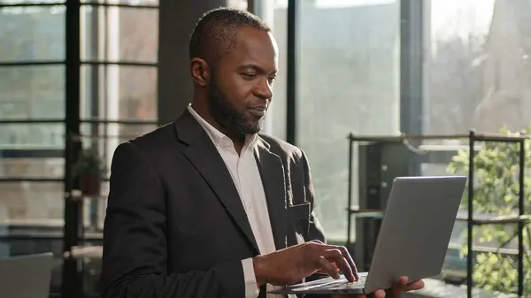 African American multiethnic businessman employee professional manager using laptop typing email on keyboard focused mature senior man search digital data stand with computer in sunny office company