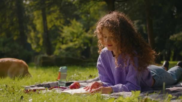 若い白人少女 都市公園の芝生の森で本を読んでいる女性 休憩週末日当たりの良いレジャー リラックス ドッグ小さな子犬のウェールズのコルギとリラックスしてアウトドアをリラックス — ストック動画