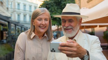 Mutlu gülümseyen Kafkasyalı yaşlı bir çift selfie çekerek gülerek konuşan bir alet cep telefonuyla şehir dışında geziyor. Yaşlı kadın gülümsüyor. Sosyal medya videosu dışarıda online video bloğu arıyor.