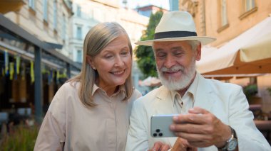 Mutlu gülümseyen Kafkasyalı yaşlı bir çift selfie çekerek gülerek konuşan bir alet cep telefonuyla şehir dışında geziyor. Yaşlı kadın gülümsüyor. Sosyal medya videosu dışarıda online video bloğu arıyor.