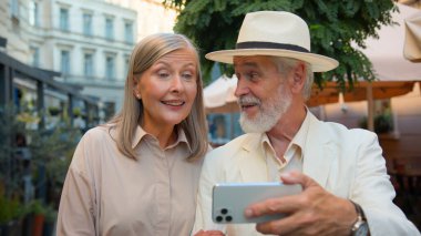 Mutlu büyükanne ve büyükbaba blogları. Yaşlı çifti kapatın. Emekli aile, beyaz kadın... Alet cep telefonuyla konuşuyor... Tatil günlerini şehir merkezindeki kafenin dışında internet günlüğünde konuşuyorlar.