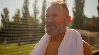 Kafkas Avrupalı, boynu havluyla yaşlı bir adam sıkı spor antrenmanından sonra şehir stadyumunda yürüyor sağlık hizmetleri yaşam tarzı aktif sağIık enerjisi dinlenmesi