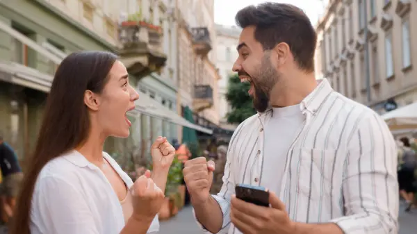 Caucasian happy couple enjoy excited man woman shocked lucky success victory mobile phone smartphone reading message good news amazed surprised open mouth city outdoors urban gen z win wonder awesome
