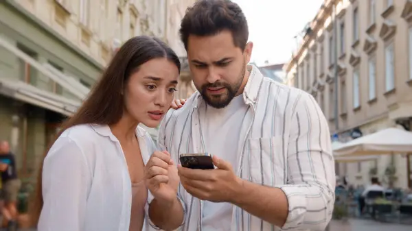 Caucasian young couple upset bad news mobile phone failure loss disappointed man woman city outside reading message street frustrated problem trouble stressed together looking at gadget screen worried